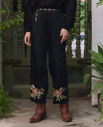 A person wearing THE GREAT. PF25 The Cuffed Pant in black with bouquet floral embroidery, styled with a black long-sleeve top, silver belt, and brown boots stands in front of a rustic green door surrounded by plants.