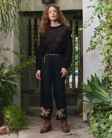 A person with curly hair stands by a green gate, wearing THE GREAT. PF25's The Cuffed Pant in black with bouquet floral embroidery, a black lace top, belt, and brown boots amid lush green plants.