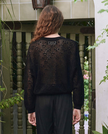 A person with long brown hair stands outdoors, facing away, wearing THE GREAT. PF25 The Crochet Pullover in black and black pants. Green foliage and wooden railings are seen in the background.