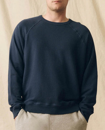 A person in THE GREAT. FALL 23 MEN The Men's College Sweatshirt -- True Navy, made of 100% cotton, pairs it with light pants and stands before a beige fabric backdrop.