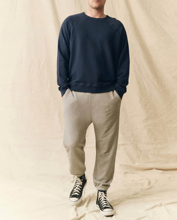 A person stands against a beige backdrop wearing THE GREAT. FALL 23 MEN The Men's College Sweatshirt in True Navy, made from 100% cotton, paired with light gray joggers and black high-top sneakers.