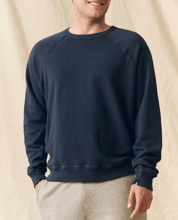 A person wears THE GREAT. FALL 23 MEN The Men's College Sweatshirt in True Navy (100% cotton) and light gray sweatpants, standing against a beige background.