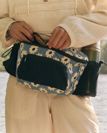 A person in a beige outfit adjusts their stylish Adventure Hip Pack - Falling Daisy Multi by THE GREAT. SP25 TGO, complete with a green water bottle nestled in the mesh pocket, ready for adventure by the tranquil lakeside.