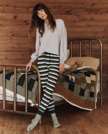 A person stands by a metal bed wearing a cozy The Sleep Cut Off Sweatshirt in Light Heather Grey from THE GREAT. HOL 24 SLEEP, alongside striped pants and socks. The bed is adorned with a patchwork quilt and topped with a teddy bear, while the room's wooden walls establish an ideal cold-weather set ambiance.