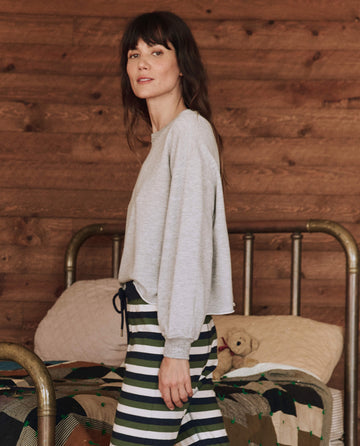 In a room adorned with a wooden wall and an old-fashioned metal bed, a person enjoys the cozy warmth of The Sleep Cut Off Sweatshirt in Light Heather Grey by THE GREAT. HOL 24 SLEEP, paired with striped pants—a perfect cold-weather set.