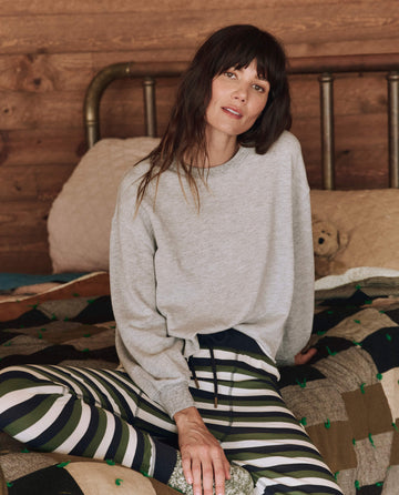 A person relaxes on a bed, wrapped in a quilted blanket, wearing a cozy Light Heather Grey Sleep Cut Off Sweatshirt by THE GREAT. HOL 24 SLEEP paired with striped pants, all set against a wooden wall backdrop—ideal for cold-weather comfort.