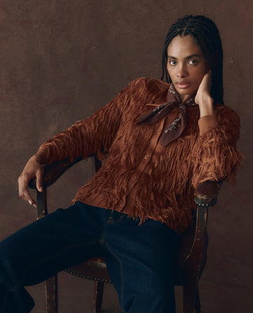 A person with braided hair sits in a wooden chair, wearing THE GREAT. F25 D1 The Fringe Cardigan in Bronze, styled with a brown scarf and dark jeans, looking directly at the camera.