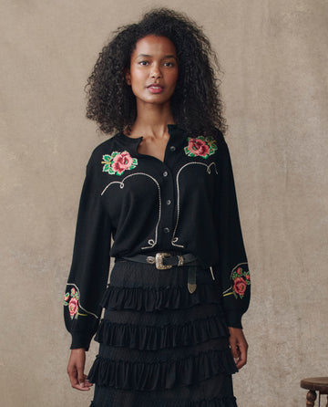 A woman stands against a beige background wearing THE GREAT. H25 COLLECTION's The Heirloom Rose Western Cardigan in black, paired with a black tiered skirt and belt.