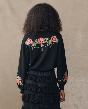 A person with curly hair faces away, wearing THE GREAT. H25 COLLECTION's The Heirloom Rose Western Cardigan in black, featuring pink floral embroidery, paired with a black layered ruffle skirt.