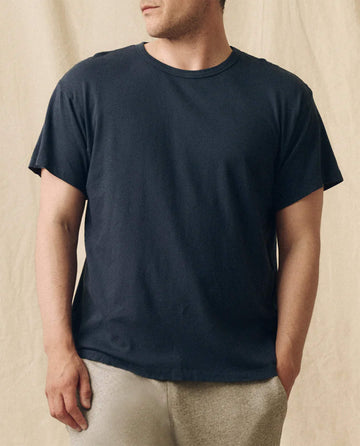 A man wears THE GREAT. FALL 23 MEN The Men's Slim Tee in True Navy with light-colored pants, standing before a beige fabric backdrop.