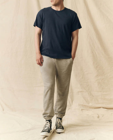A man stands against a beige fabric backdrop, wearing THE GREAT. FALL 23 MEN The Men's Slim Tee in True Navy, light gray sweatpants, and black high-top sneakers with his hands in his pockets.
