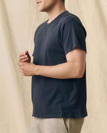 A person wears THE GREAT. FALL 23 MEN The Men's Slim Tee in True Navy, paired with light gray pants and shown from the side against a beige fabric backdrop. Only the upper body of this 100% cotton vintage t-shirt is visible.