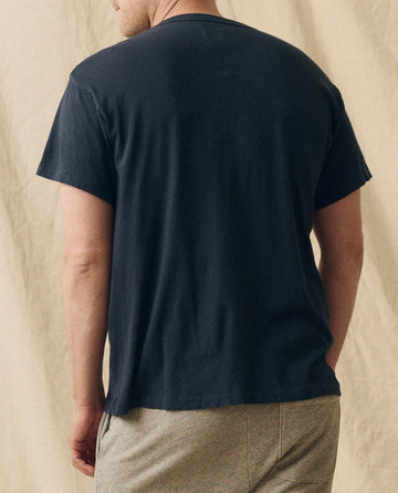 A man stands with his back to the camera, wearing THE GREAT. FALL 23 MEN The Men's Slim Tee in True Navy and light-colored shorts, set against a neutral fabric backdrop.
