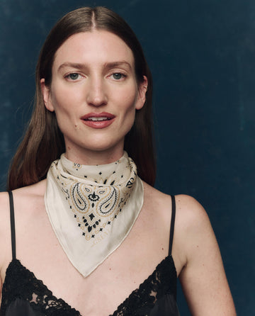 A woman with straight brown hair poses against a dark blue background, wearing a black lace top and The Silk Bandana in Cream by THE GREAT. F25 COLLECTORS around her neck.