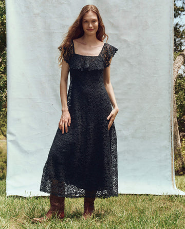 A woman in a flowing The Balcony Dress -- Navy by THE GREAT. PS25 and boots stands on the grass, smiling, with a light-colored backdrop and lush greenery in the background.