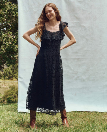 A woman stands outdoors on grass in a navy dress named "The Balcony Dress" by THE GREAT. PS25, framed by a white backdrop and trees, smiling with her hands on her hips.