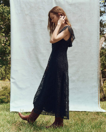 A person stands gracefully outdoors in THE GREAT. PS25's The Balcony Dress in navy, complemented by brown boots against a light backdrop.