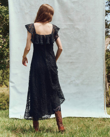 A woman in The Balcony Dress by THE GREAT. PS25, navy lace, paired with brown boots, stands on grass facing away. She gazes thoughtfully at the picturesque outdoor backdrop.