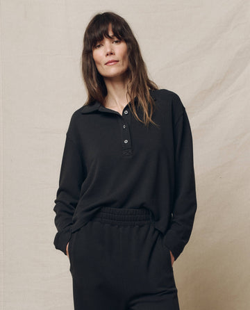 Someone with long hair stands against a plain backdrop, wearing THE GREAT. SP25 POLO LOUNGE's The Rugby Sweatshirt in Almost Black with matching pants.