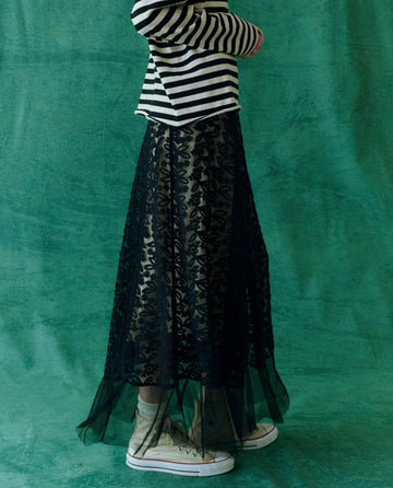 A figure in chic women's clothing dons The Era Skirt in black by THE GREAT. HOL 24, paired with a striped shirt and white high-top sneakers, standing out against a vibrant green background. This look effortlessly captures the essence of modern fashion.