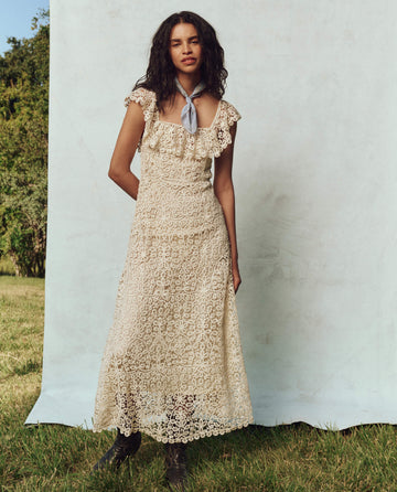A person stands outdoors in The Balcony Dress, a cream lace design by THE GREAT. PS25, paired with a blue bandana around their neck.