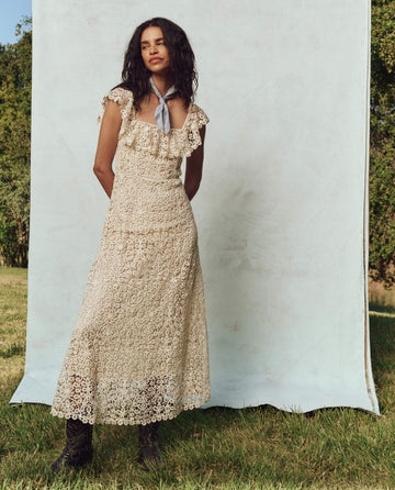 Wearing The Balcony Dress in cream, a person stands on the balcony wrapped in a scarf, surrounded by lush greenery under gentle lighting. Brand: THE GREAT. PS25.