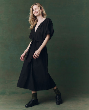 A person with long blonde hair wears THE GREAT. F25 D2 Fairground Dress in black, featuring puffed sleeves and a belt, paired with dark socks and black shoes, posing against a green background.