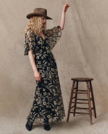 A woman in a brown hat and The Festal Dress -- Brocade Floral by THE GREAT. H25 COLLECTION stands beside a wooden stool against a plain beige backdrop.