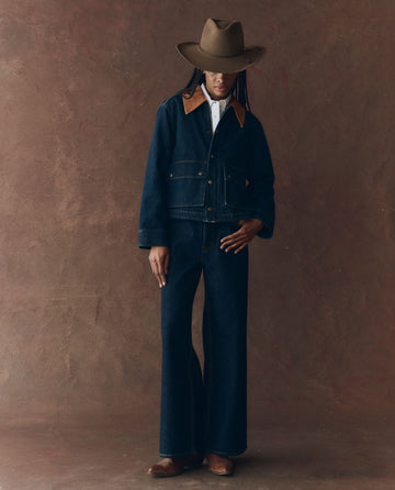A person in THE GREAT. F25 D1 The Denim Swingy Jacket in Midnight Rinse, a white shirt, wide-leg denim jeans, cowboy hat, and brown boots stands against a brown background.