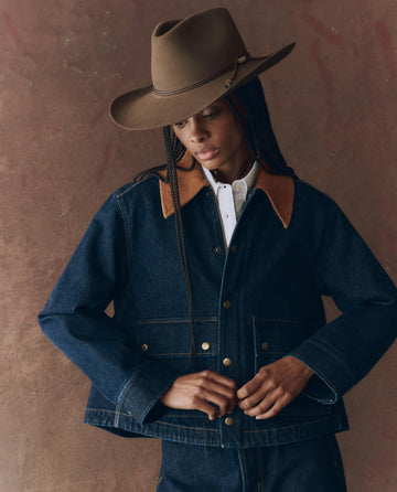 Person wearing THE GREAT. F25 D1 The Denim Swingy Jacket in Midnight Rinse, paired with a wide-brimmed brown hat and a white shirt, standing against a brown textured background.