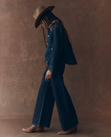 A person in profile walks against a brown textured background, wearing THE GREAT. F25 D1's The Denim Swingy Jacket in Midnight Rinse.