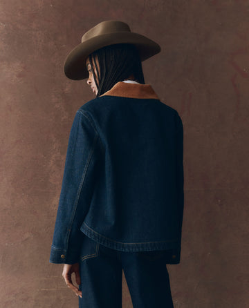 A person faces away from the camera against a brown textured background, wearing THE GREAT. F25 D1’s The Denim Swingy Jacket in Midnight Rinse and matching denim pants, styled with a wide-brimmed hat and brown-collared details.