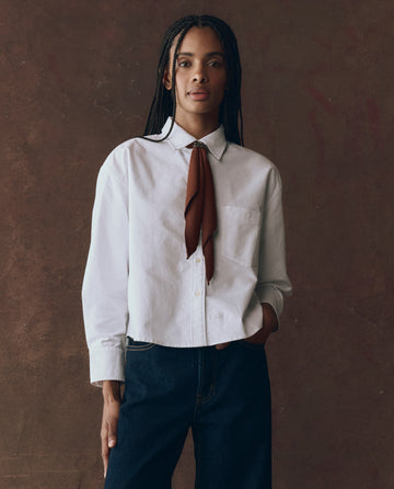 A person stands against a brown background wearing THE GREAT. F25 D1 The Cropped Oxford Shirt in white, dark jeans, and a brown necktie, with one hand in their pocket.