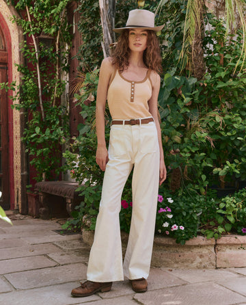 A woman stands outdoors on a stone path surrounded by greenery and flowers, wearing THE GREAT. PF25 The Henley Slim Tank in Bronze Stripe, a beige hat, white pants, and brown shoes.