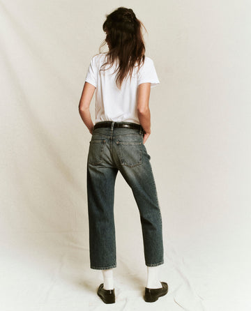 A person with long hair is standing with their back to the camera, wearing The Slim Tee in True White from THE GREAT. CORE KNITS, along with blue jeans, white socks, and black shoes. The plain, light-colored background subtly highlights the 100% cotton fabric of the shirt.