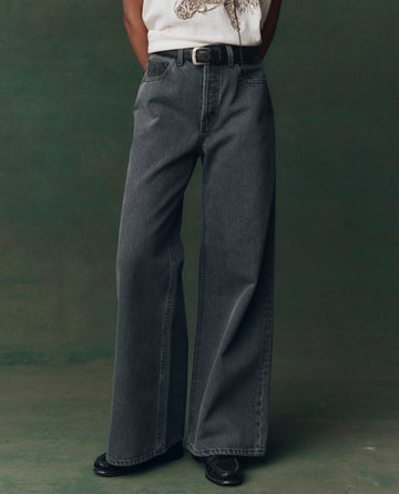Person wears THE GREAT. F25 D2 The Slouch Wide Leg Jean in Graphite Wash, paired with a black belt, white graphic tee, and black shoes, standing against a green backdrop.
