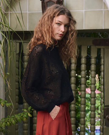 A woman with wavy hair stands outdoors by a wooden gate with greenery, wearing THE GREAT. PF25 The Crochet Pullover in black and rust-colored pants.
