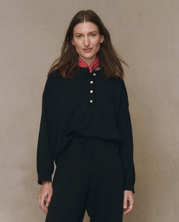 A woman wears THE GREAT. F25 HIGH TWIST The Henley Sweater in black with matching pants, layered over a red patterned collar for a vintage menswear touch, standing against a simple beige background and facing the camera.