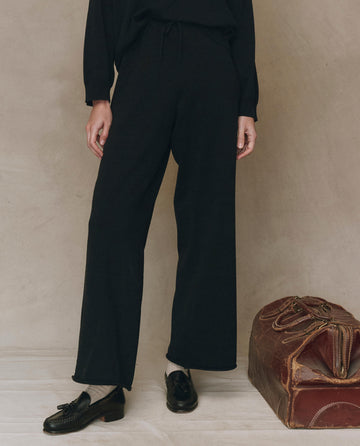 A person stands against a neutral wall wearing THE GREAT. F25 HIGH TWIST The Rec Sweater Pant in black with black loafers; a brown leather bag rests on the floor beside them.