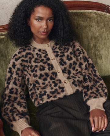 A person with curly hair sits on a green sofa, wearing THE GREAT. H25 COLLECTION’s The Windsor Cardigan in leopard print and dark pinstriped pants.