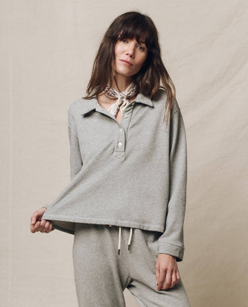 A woman in THE GREAT.'s Varsity Grey Rugby Sweatshirt holds the hem with one hand. Her long hair cascades around a scarf tied at her neck, set against a neutral background.
