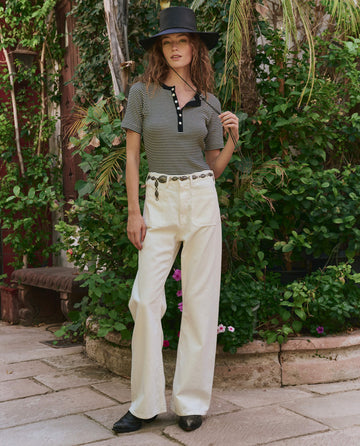 A woman stands outdoors in front of lush greenery, wearing THE GREAT. PF25 Baseline Henley in Black & Cream Stripe, high-waisted white pants, black shoes, and a black wide-brim hat for an effortless casual look.