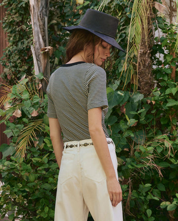 A person wearing THE GREAT. PF25 The Baseline Henley in Black & Cream Stripe, a black hat, and white pants stands outdoors with their back to the camera, surrounded by green foliage.