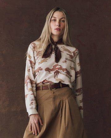 A woman with long blonde hair wears THE GREAT. The Slouch Sweatshirt in the Riding Horses Print, styled with a brown scarf and belted tan skirt, standing against a brown background.