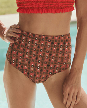 A person wearing THE GREAT. SP25 SWIM's The Reversible Mid Rise Brief in Rust Tile Print and Hot Red with a red ruffled bikini top stands near a swimming pool.