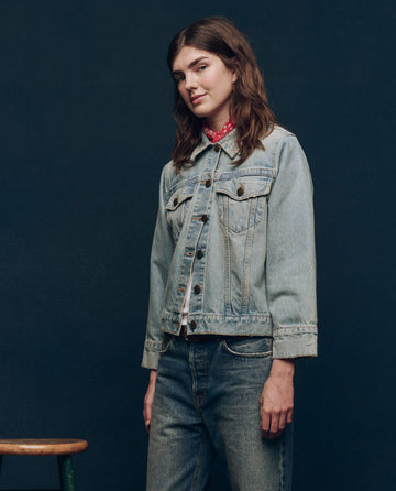 A person wearing The Slouchy Jean Jacket in Great Plains Wash by THE GREAT. SP25 COLLECTORS stands against a dark background, with a red bandana under the collar and part of a green stool visible to the left.