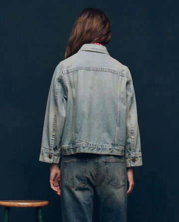 A person wears The Slouchy Jean Jacket in Great Plains Wash by THE GREAT. SP25 COLLECTORS, with jeans, facing away from the camera against a dark backdrop near a wooden stool. The vintage-inspired jacket exudes effortless Made-in-LA charm.