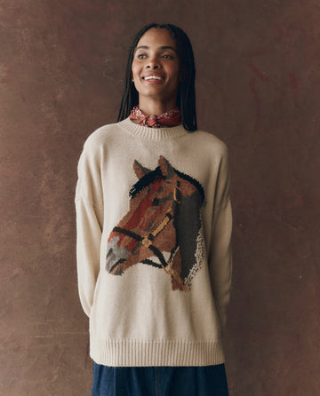 A person wearing THE GREAT. F25 D1 The Stable Pullover in Natural, featuring a bold horse-head intarsia, stands smiling slightly in front of a brown background.