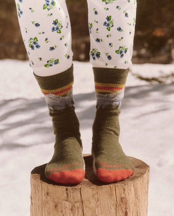 A person stands on a tree stump wearing white leggings with blue flowers and The Ridge Runner Socks in Evergreen, embracing the snowy outdoor setting in their luxurious merino wool attire by THE GREAT. FALL 23 TGO.