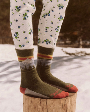 Person wearing patterned thermal leggings and The Ridge Runner Sock in Evergreen, standing on a wooden stump in a snowy outdoor setting, ready for their everyday adventure.